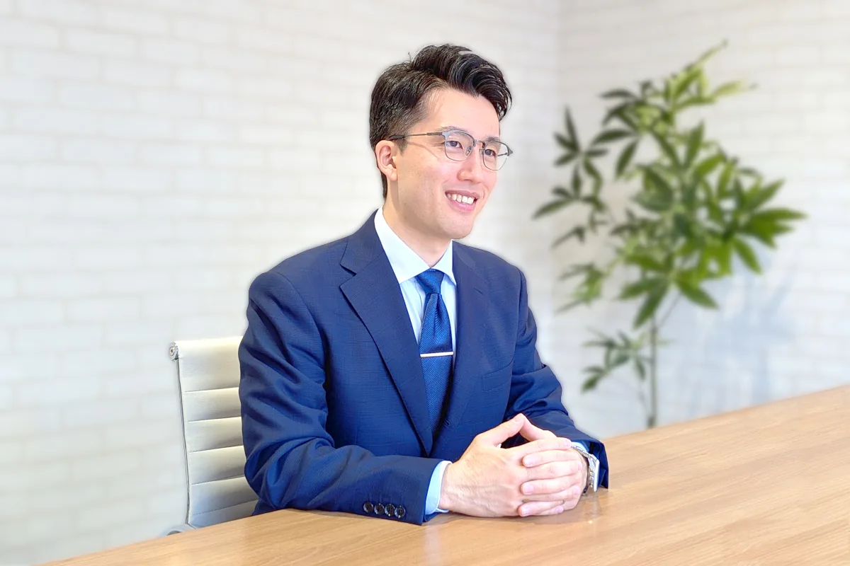 白いレンガ調のオフィスにて木目のテーブルを前に穏やかな笑顔で話を聞く代表の相田暁文