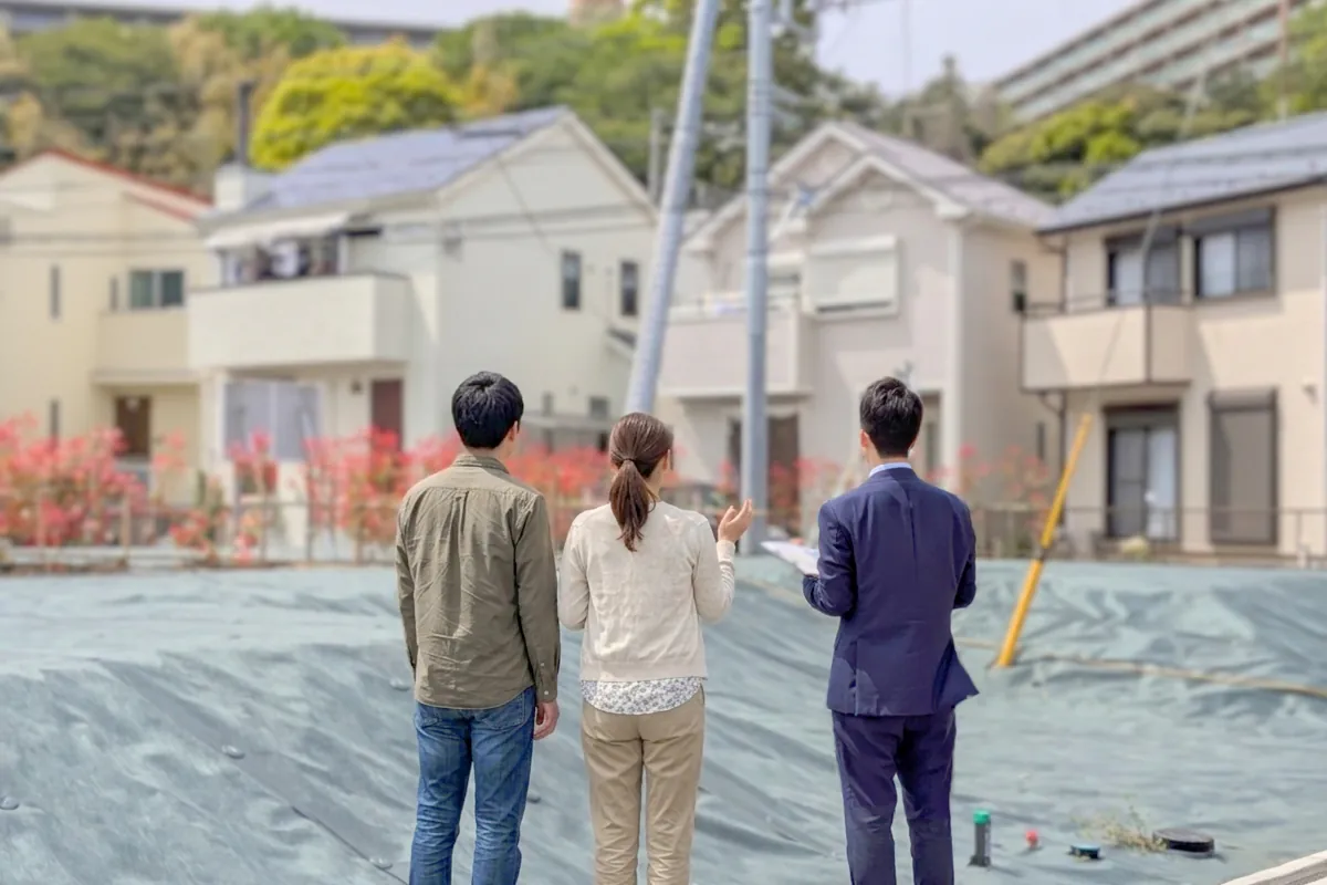 防草シートが敷かれた更地にて紺色のスーツを着た専門家（相田暁文）が手に持つ資料を使ってお客様ご夫婦に説明している様子